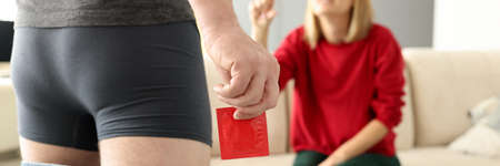 Man in shorts pulling jeans down stands with condom in hand close-up in front of girl. Plastic...