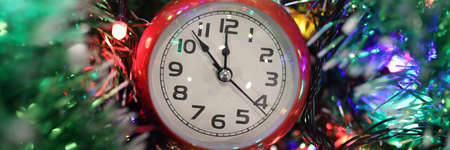 Red alarm clock lying on table in bright garland and tinsel closeup. Crown of new year and christmas...