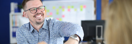 Cheerful man in glasses show client watch on his hand at workplace. Visitor sit opposite employee.