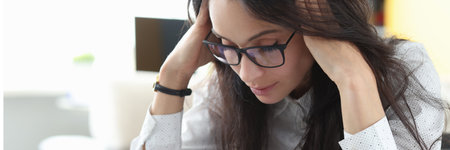 Tired woman with glasses sit at table and massages her head with her hands and think. Employee...