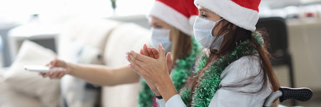 Woman in red cap and jewelry around her neck in wheelchair and face shield claps hands with her friend with tv remote control. Celebrating new year and christmas at home self isolation concept.の写真素材