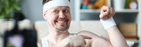 Joyful young man in sport headband looking at camera and smiling while demonstrating biceps muscle