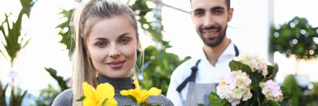 Charming young woman and bearded man in apron looking at camera and smiling while demonstrating flowers in plant storeの写真素材
