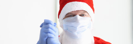 Man in santa claus costume and protective medical mask and gloves stands with his hands together and smiles. Safe New Year and Christmas celebration in coronavirus pandemic conceptの写真素材