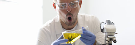 Male laboratory technician looks in surprise at test tube of golden liquid with marijuana symbol....