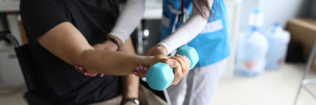 Paralyzed man sit in wheelchair and hold blue dumbbell on his outstretched arm. Femail doctor help patient to knead his hand in clinic.の写真素材