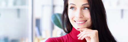 Beautiful young brunette in red smiles and looks out the window. Office worker distracted from workの写真素材