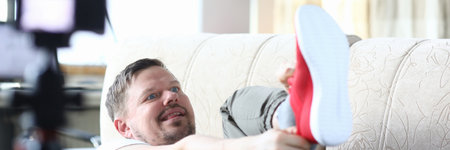 The guy lies on the couch and pulls red sneakers. A man in his apartment is reviewing new sports shoes. Video blog about buying shoesの写真素材