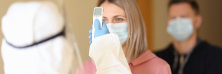 Doctor in protective suit measuring temperature of people in line with non-contact infrared...
