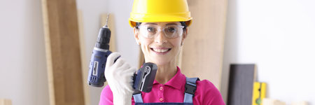 Woman builder in overalls and protective helmet holding drill in hands in workshop. Male professions for women conceptの写真素材