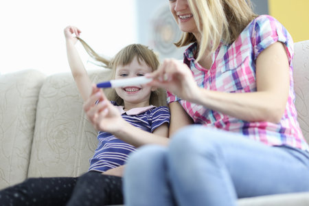Joyful mom and daughter are sitting on couch and holding pregnancy test. How to prepare child for new addition to the family conceptの写真素材