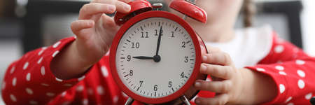 Little girl in background presses button of red alarm clock with her hand in morning close-up. Organization of time in preschool educational institutions.の写真素材