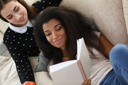 Two women lying on couch and reading book. Female friendship and relationship conceptの写真素材