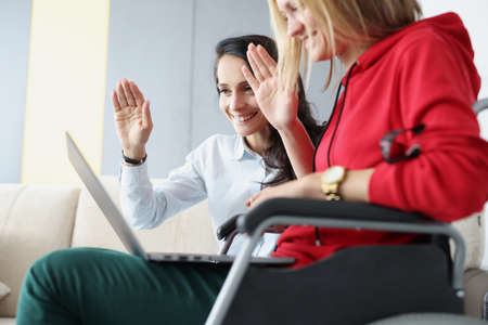 Woman with friend sitting in wheelchair and holding laptop waving to monitor. Remote work for people with disabilities conceptの写真素材