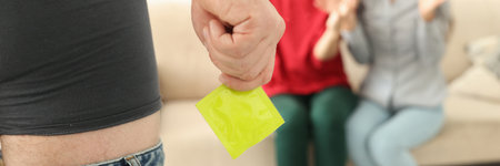 Man stands in shorts with his pants down and holds condom in front of two women. Relationship in...