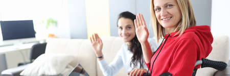 Young disabled woman in wheelchair with helper is sitting by laptop screen and waving hello. Social...