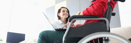 Disabled woman in wheelchair communicates with smiling friend with laptop on her lap. Psychological assistance to people with disabilities concept.の写真素材