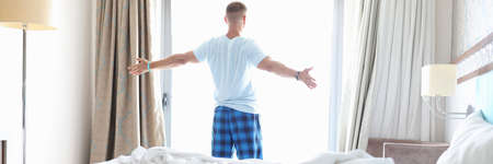 Man stand with his back near window with his arms outstretched to sides. Morning awakening in hotel room.の写真素材