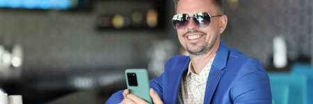 Businessman hold phone in his hand. Happy man in blue jacket with beard and sunglasses sit at bar...