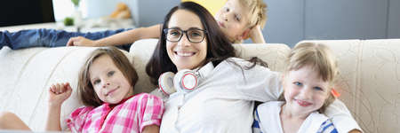 Woman is sitting on sofa with two girls and a boy is standing next to laptop. Teaching children...