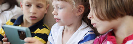 Three children are sitting on the couch and looking at smartphone screen. How to choose a smartphone...