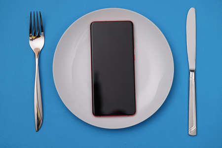 Mobile phone lying in white plate near fork and knife. Phone addiction concept