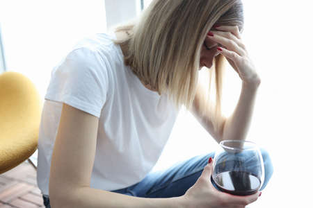 Depressed woman sits with her head bowed and holds glass of wineの写真素材