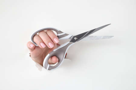 Female hand holds metal scissors in hole made of white paperの写真素材