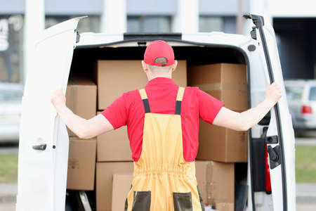 Male courier stands behind car with open doors in cardboard box showroom