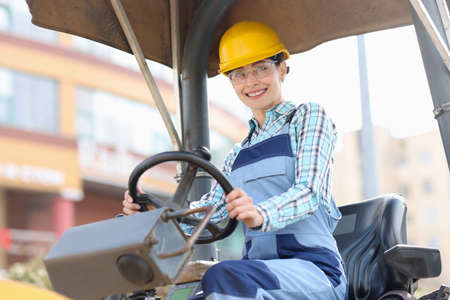 Young woman builder in hard hat sitting behind wheel of asphalt paver