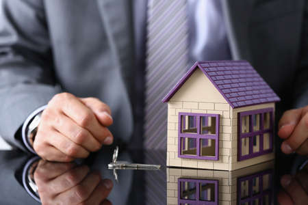 Businessman sitting at table with toy wooden house and metal keys closeupの写真素材