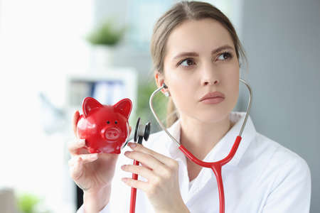female doctor holds red pig piggy bank and applies stethoscope to itの写真素材