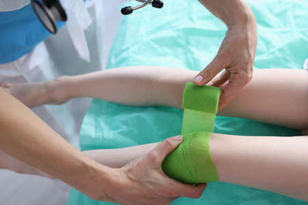Doctor is bandaging child sore leg with green bandage in clinic closeupの写真素材