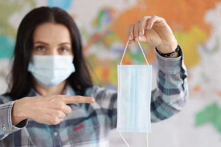 Woman in protective medical mask points at medical mask with her finger against background of world...