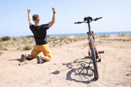 Rear view of happy excited winner traveler kneel down with raised hands, next to him is bicycleの写真素材