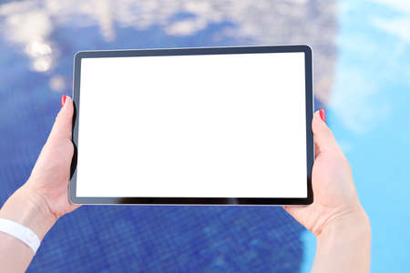 Female hands hold tablet with blank white screen above pool water