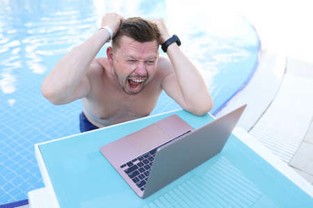 Man screaming and holding his hair and looking at laptop screen on side of poolの写真素材