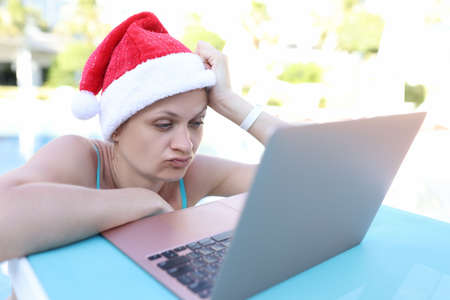 Tired woman in santa claus hat looking at laptop screen on side of poolの写真素材
