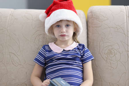 Little sad girl sitting on sofa wearing santa claus hatの写真素材