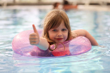 Little girl swimming in pool using inflatable circle and showing thumb upの写真素材