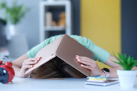 Tired in stress woman hides her head with laptop at workplace.