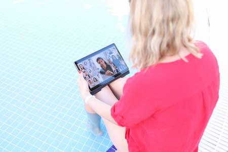 Young woman is holding online business conference via tablet while sitting on edge of pool