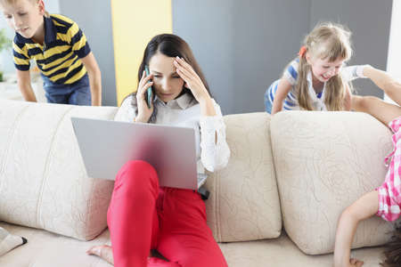 Sad young woman sitting on sofa with laptop and holding her head near children playing at homeの写真素材