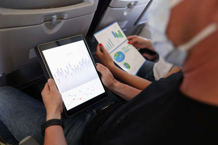 Man and woman are flying in airplane wearing protective masks with digital tablet and documents in...