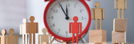Wooden figurines with one red figure stand next to alarm clock