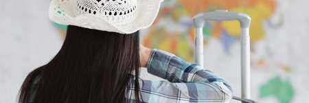 Woman sits with her back in white hat and suitcase and looks at map of worldの写真素材