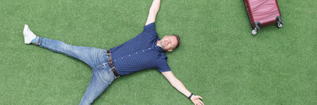 Happy man lies on green lawn with suitcase top viewの写真素材