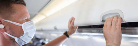 Man in protective medical mask closing carry-on luggage rack in airplaneの写真素材