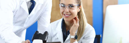 Young man and woman in white uniforms sitting behind table with microscopeの写真素材