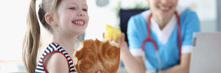 Smiling little girl eats bun on background of doctorの写真素材